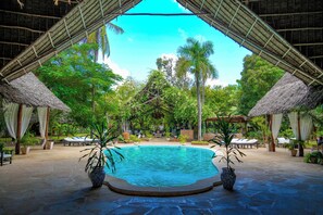 Un área de piscina con una piscina azul cristalina, rodeada de sillones de descanso y plantas en macetas, bajo una estructura de techo de paja.