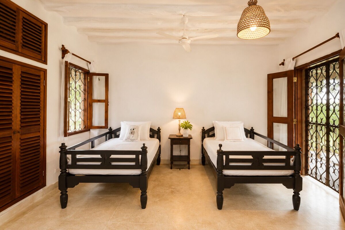 A room with two beds, a ceiling fan, a lamp, and a window with a decorative screen.