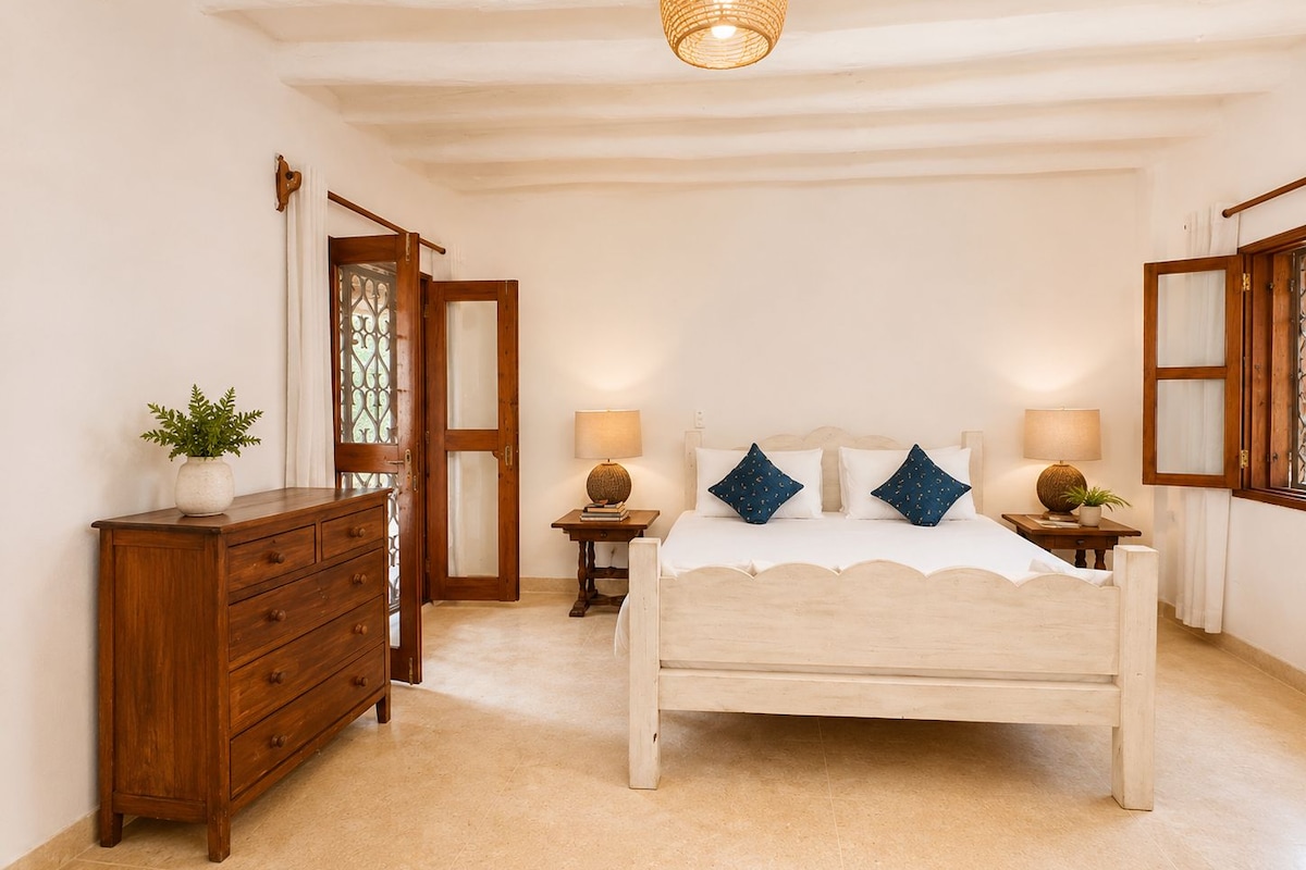 Un dormitorio con cama, cómoda de madera, dos mesitas de noche con lámparas, una lámpara colgante y una puerta con panel de vidrio.