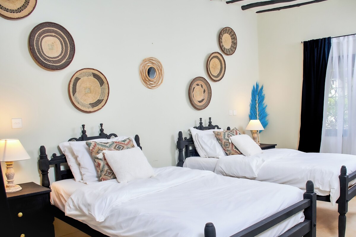 A hotel room with two beds, a nightstand with a lamp, and decorative woven wall hangings.