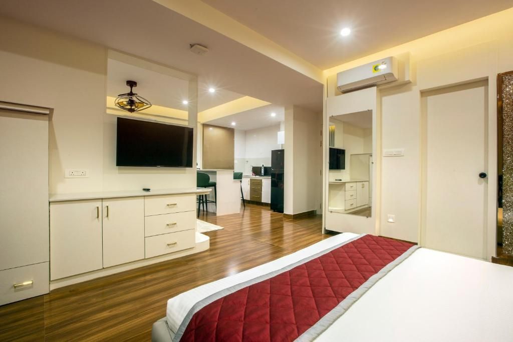 A modern hotel room with a large bed, a flat-screen TV mounted above the bed, a kitchenette area, and a bathroom with a mirror and sink.