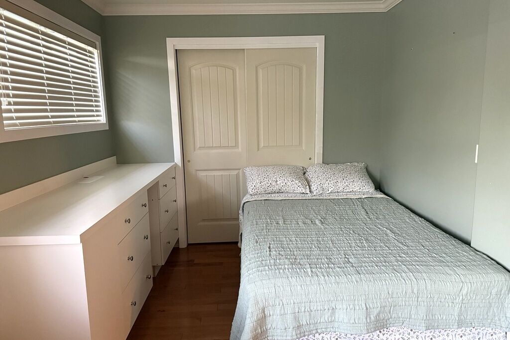 A bedroom with a bed, a dresser, a door, and a window with blinds.