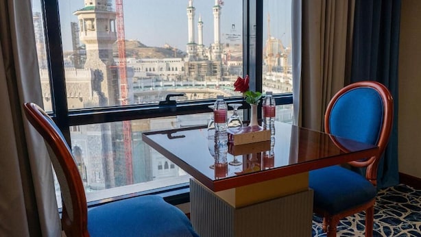 Hotelkamer met glazen tafel, twee stoelen, een vaas met een roos en uitzicht op een stadsbeeld met hoge gebouwen en een toren.