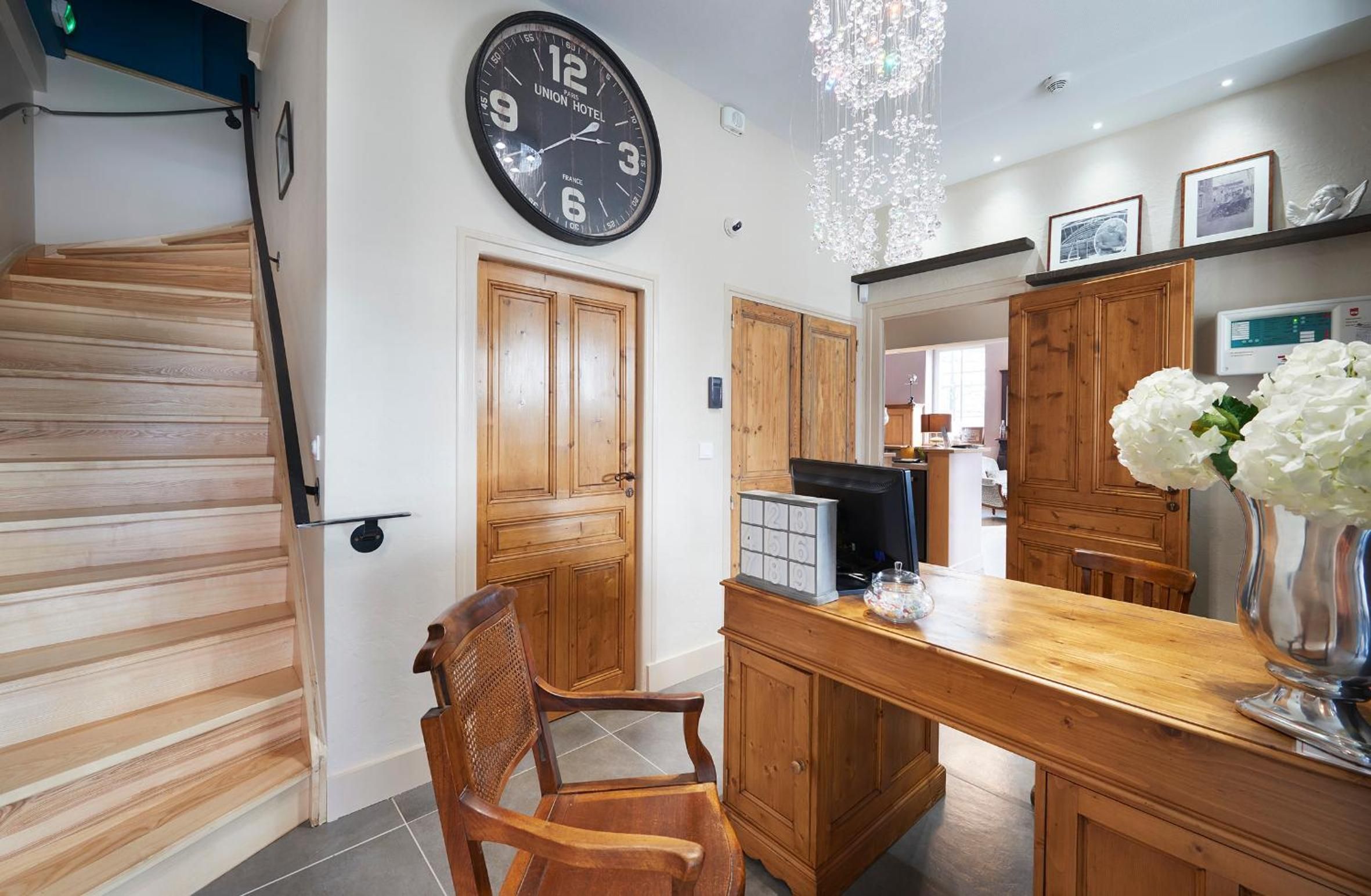 A hotel interior with a wooden staircase, a dining area with a wooden table, chairs, and a clock on the wall.