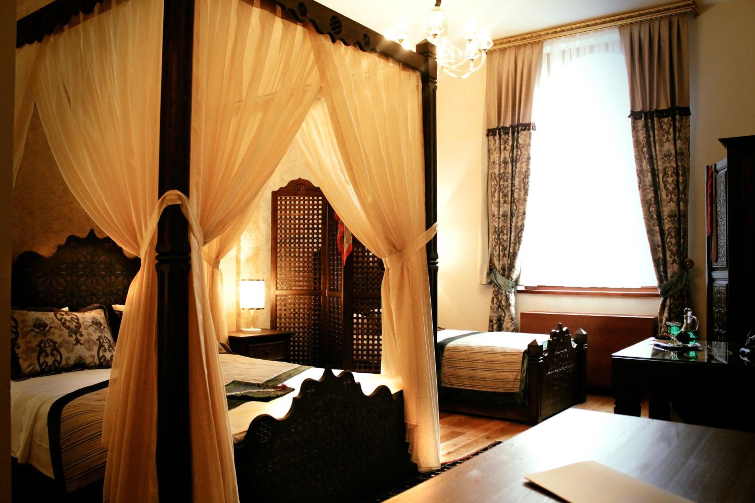 A four-poster canopy bed with a wooden headboard, a bedside table with a lamp, and a window with curtains.