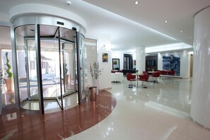 A lobby with a revolving door, a reception desk, and a seating area with red chairs. - Hotel Eridano (Sannazzaro de Burgondi)