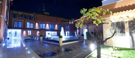 A courtyard with a fountain, potted plants, and a building with multiple windows.