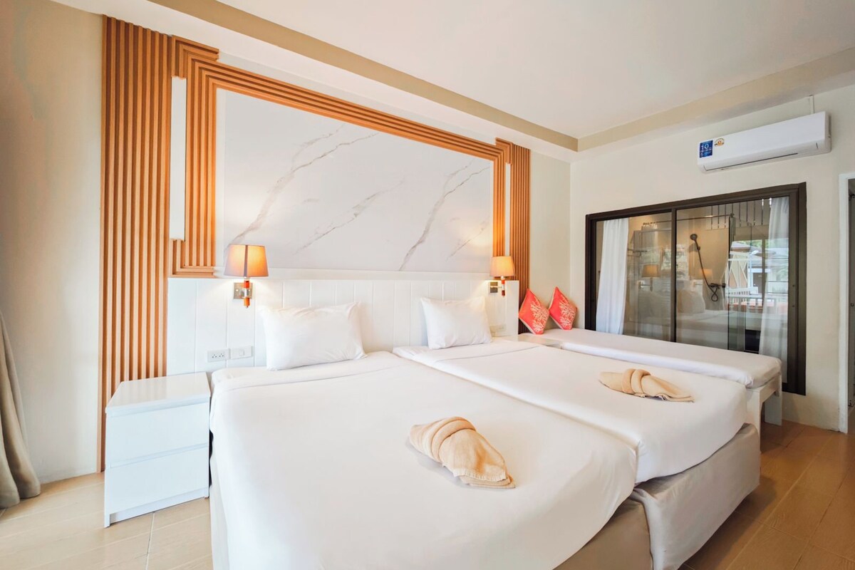 Une chambre d’hôtel avec deux lits, une tête de lit à motifs de marbre, une table de chevet blanche et une porte en verre donnant sur un balcon.