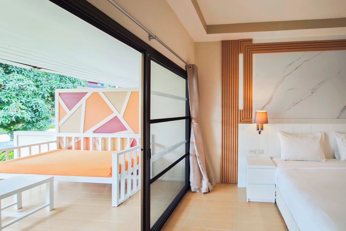 A hotel room with a bed, a bedside table, a lamp, and a view of greenery outside.
