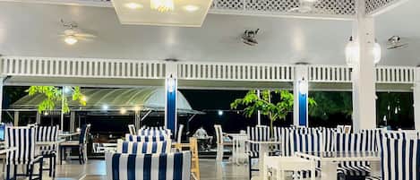 A spacious outdoor seating area with striped chairs and tables, under a covered section with decorative lattice panels and ceiling lights.