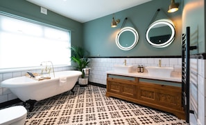A bathroom with a freestanding bathtub, two sinks, a vanity with a mirror, and patterned floor tiles. - The Bear (Bath)