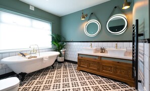A bathroom with a freestanding bathtub, two sinks, a vanity with a mirror, and patterned floor tiles. - The Bear (Bath)