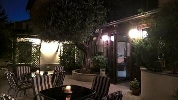 Un'area salotto esterna con tavoli e sedie, circondata da piante e un edificio con finestre illuminate.