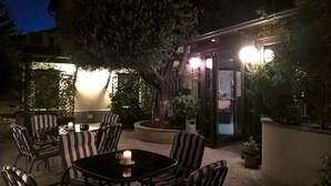 Ein Sitzbereich im Freien mit Tischen und Stühlen, umgeben von Pflanzen und einem Gebäude mit beleuchteten Fenstern.