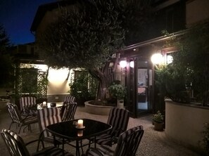 Un'area salotto esterna con tavoli e sedie, circondata da piante e un edificio con finestre illuminate.