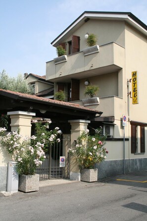 A two-story building with a yellow sign indicating a hotel. There are potted plants in front of the entrance, and a no parking sign is visible. - Hotel Rocco (Carugate)