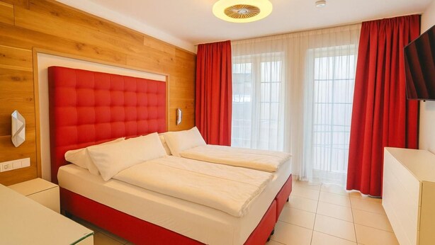 Ein Hotelzimmer mit rotem Kopfteil, zwei Betten, einer Holzwand, einem großen Fenster mit roten Vorhängen und einem Flachbildfernseher.