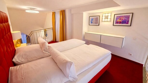 A hotel room with a double bed, a red accent wall, a staircase with yellow and white curtains, and two framed pictures on the wall. - Villa Viktoria 1 - Apartments - City Center (Ingolstadt)