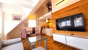Ein modernes Wohnzimmer mit einem Glastisch, violetten Sesseln, einem Flachbildfernseher, der an einer Holzwand montiert ist, und einer Erkerzone mit einer Küche.