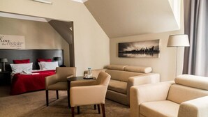 A hotel room with a bed, two armchairs, a sofa, a small table with water bottles, a cityscape painting, and a lamp.