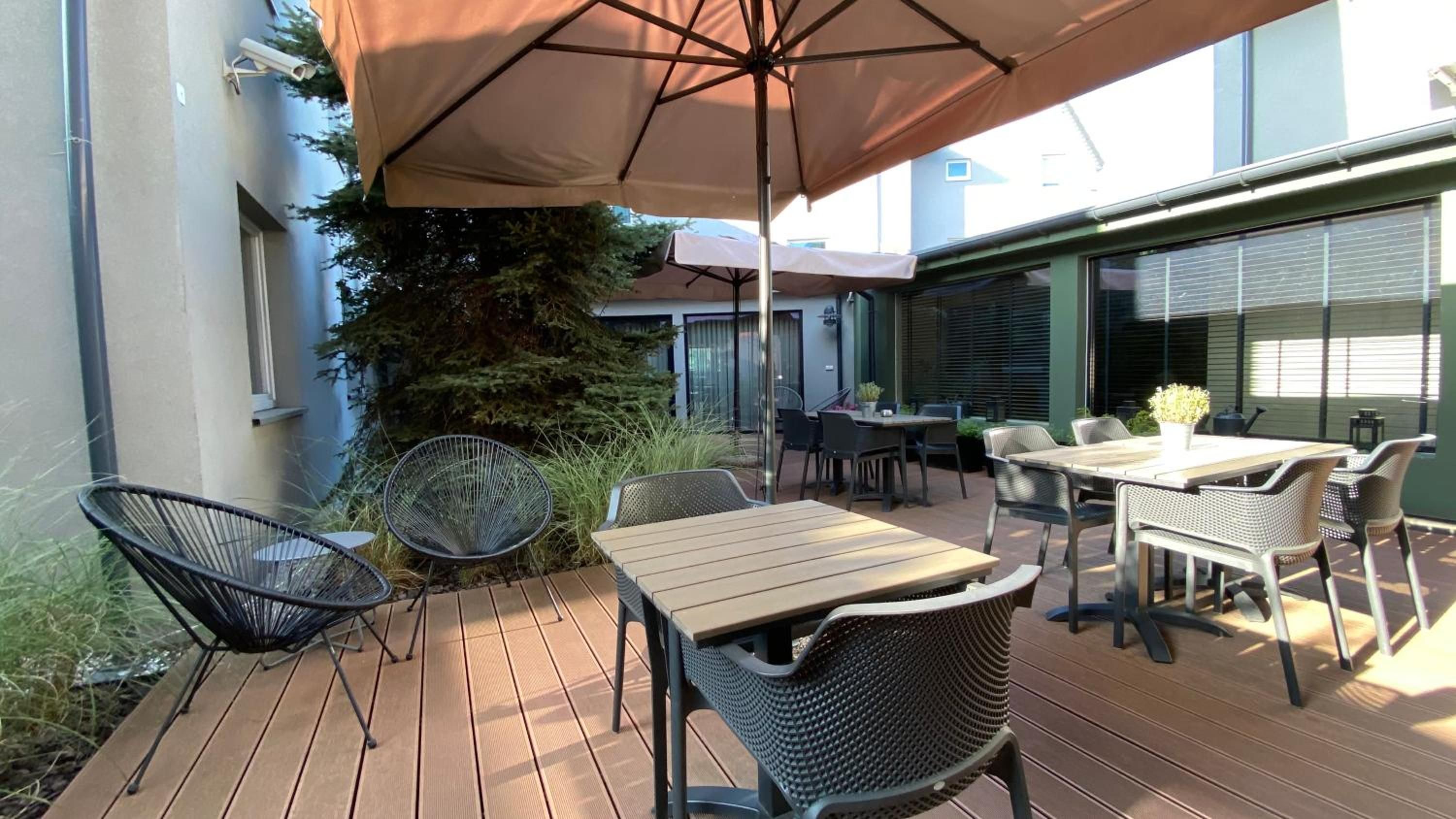 An outdoor patio with wooden tables and chairs, a large umbrella, and a potted plant.