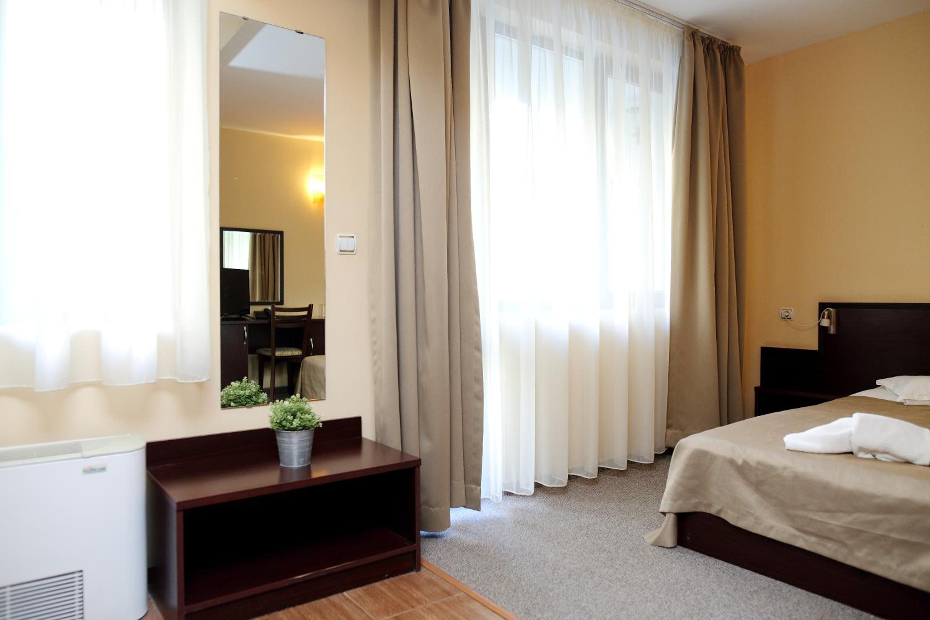 A hotel room with a bed, a desk with a chair, a mirror, and a potted plant.