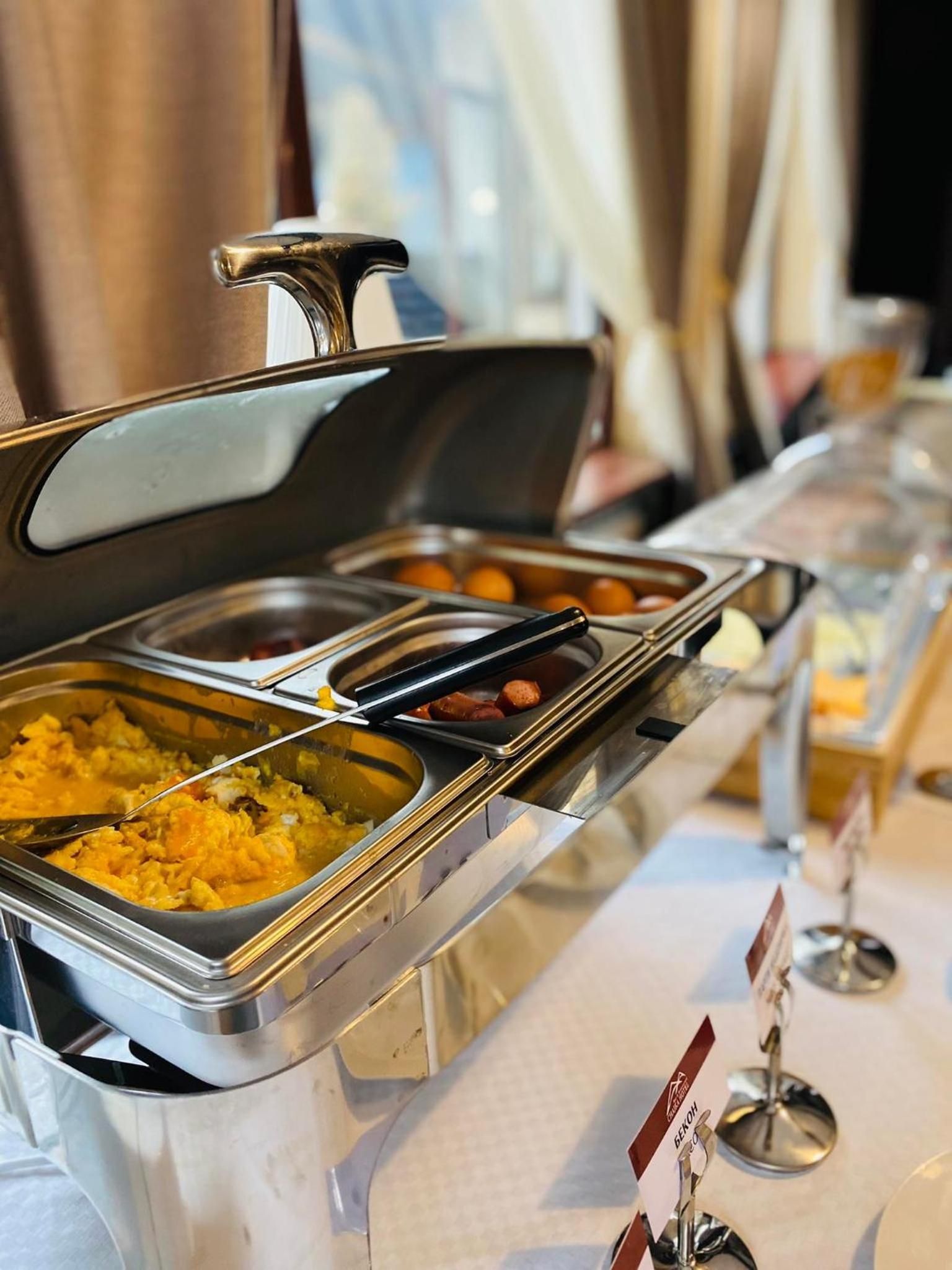 Disposição de buffet com vários recipientes de comida, incluindo ovos mexidos e salsichas.