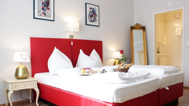 Een hotelkamer met een rode hoofdbord, witte beddengoed, een nachtkastje met een lamp en een spiegel.