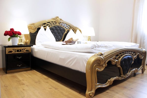 A bed with a black and gold frame, white bedding, and a nightstand with drawers and a vase of red roses. - Stadthotel Restaurant Auerhahn (Voecklabruck)