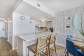 Een moderne keuken met witte kastjes, een centraal eiland en een eethoek met rieten stoelen en een blauwe tafel.