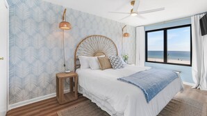 Ein Schlafzimmer mit einem Bett, einem Deckenventilator, einem Fenster mit Blick auf den Strand und Wandtapete mit Muster.