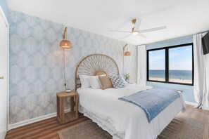 Ein Schlafzimmer mit einem Bett, einem Deckenventilator, einem Fenster mit Blick auf den Strand und Wandtapete mit Muster.