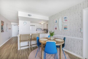 A modern kitchen with white cabinets, a microwave, and a dining area with a round table and blue chairs. - Beach Condo Loved for Location & Thoughtful Extras (Gulf Shores)