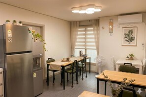 A compact kitchen with a refrigerator, a dining table with chairs, a window with curtains, and a wall-mounted clock. - Ara Guesthouse (Busan)