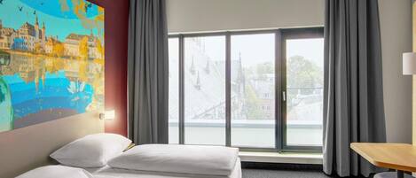 A hotel room with a bed, a desk, a chair, a large window with curtains, and a painting on the wall.