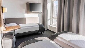 A hotel room with a bed, a TV mounted on the wall, a window with curtains, and a small table with a lamp. - B&B Hotel Zwickau (Zwickau)