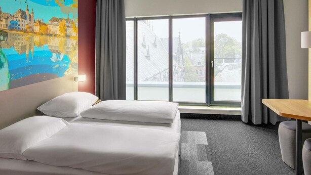 A hotel room with a bed, a desk, a chair, a large window with curtains, and a painting on the wall. - B&B Hotel Zwickau (Zwickau)