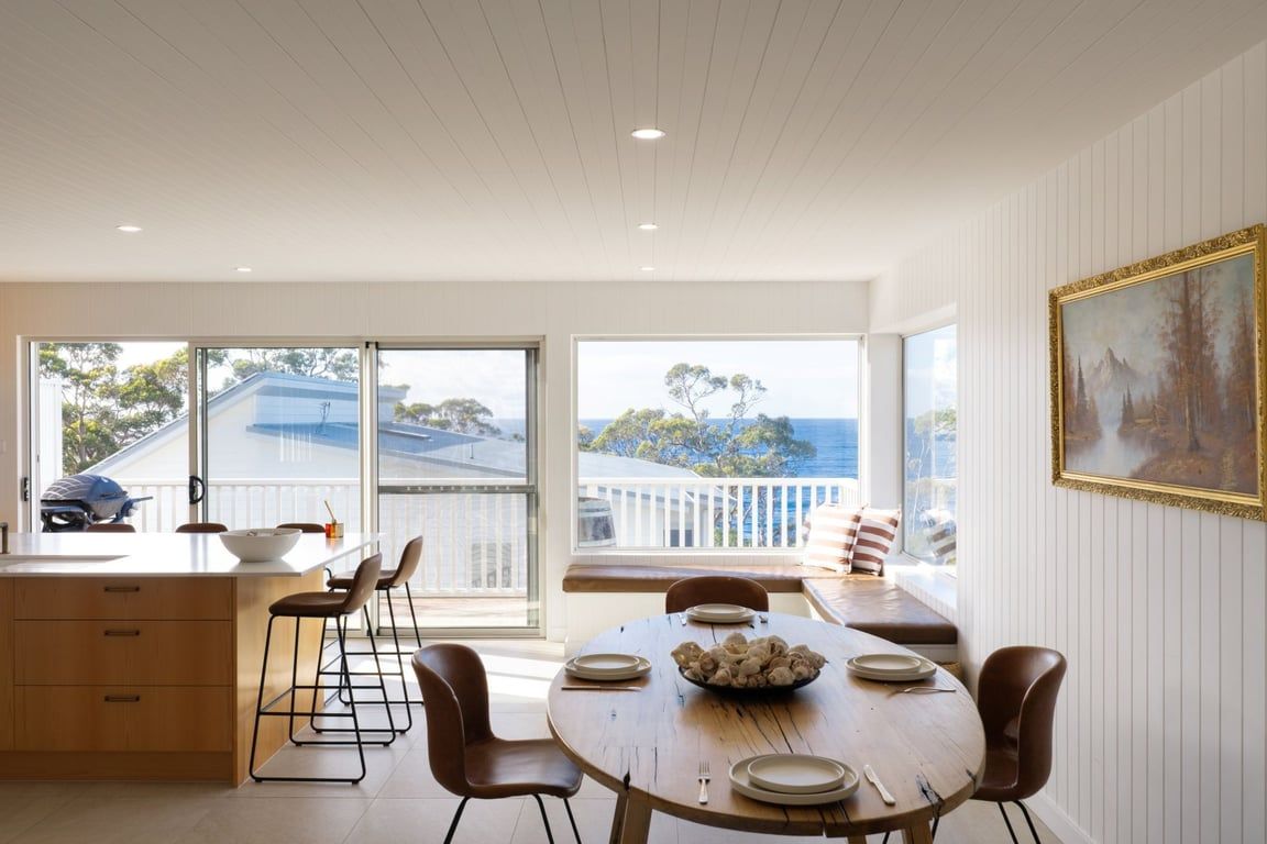 A dining area with a wooden table, chairs, and a bowl of fruit. There is a view of the sea through large windows.