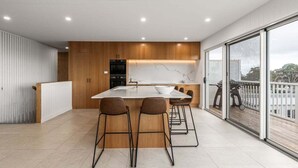 A modern kitchen with a central island, bar stools, and a view of the outdoors through large windows. - White Pebble - Bicheno Accommodation (Bicheno)