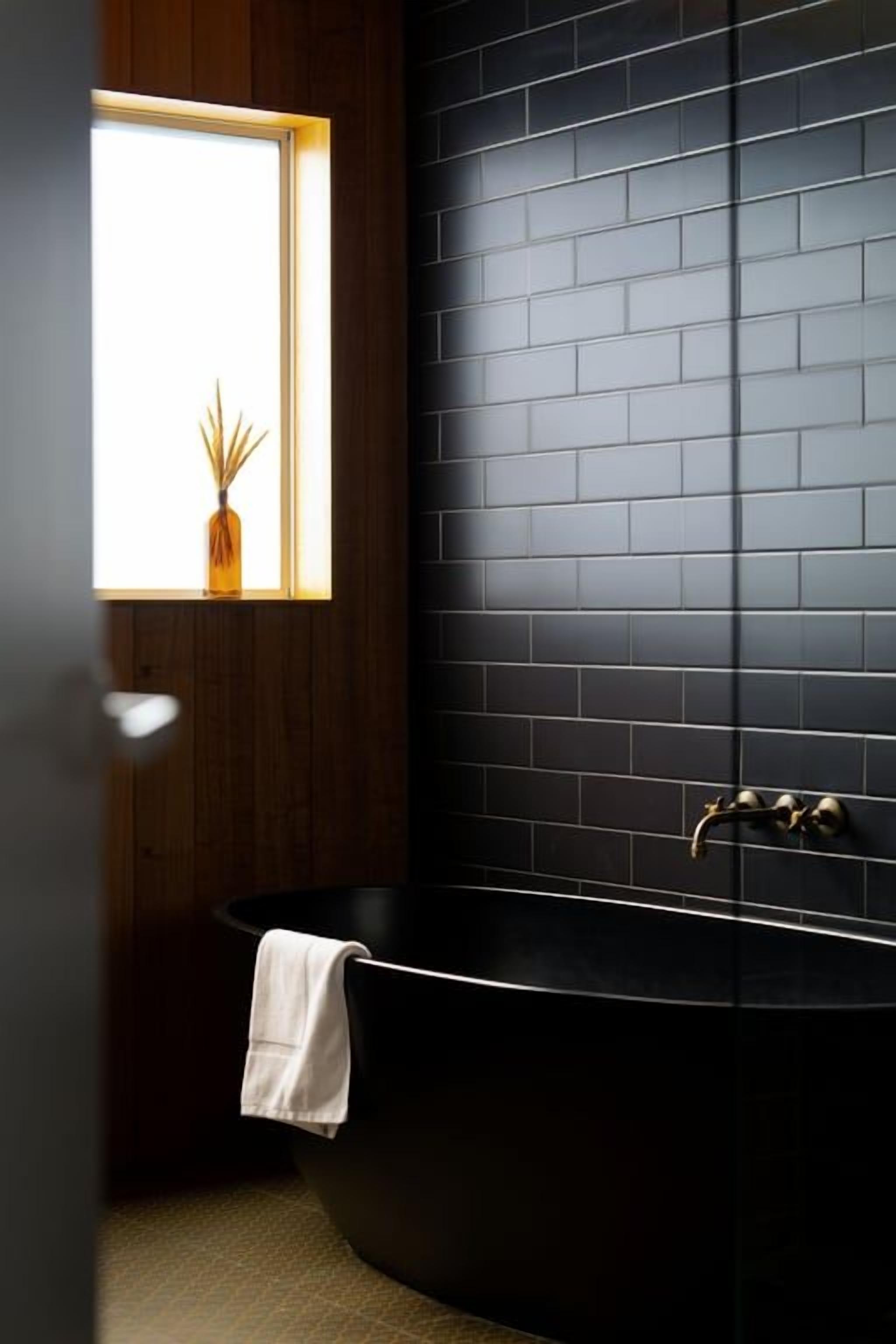 Une salle de bain moderne avec une baignoire noire, une fenêtre agrémentée d’une plante et une porte en bois.
