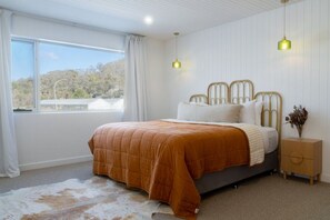 Ein Schlafzimmer mit einem großen Bett, einem Nachttisch, einem Fenster mit Vorhängen und Blick auf Bäume.