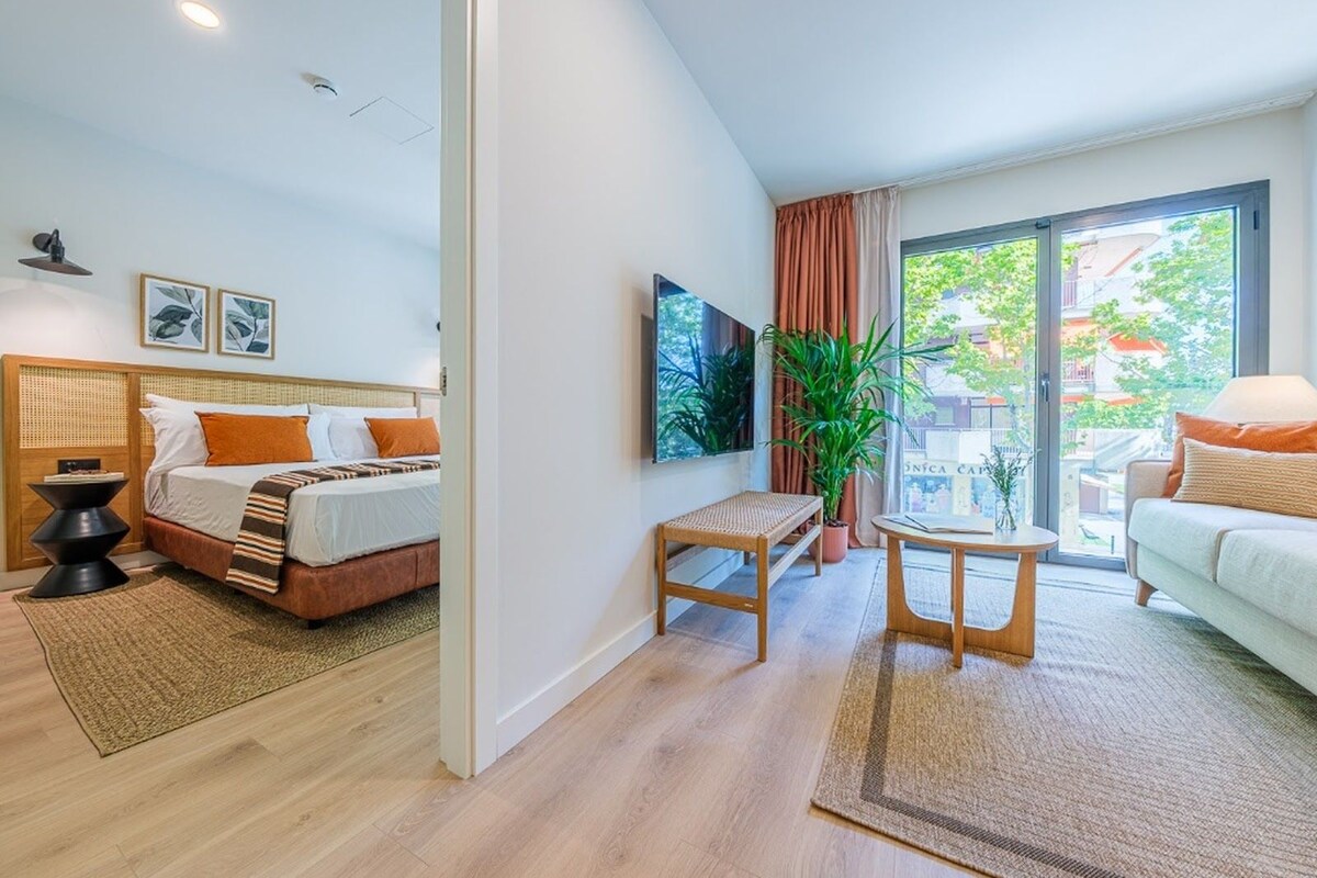 A modern hotel room with a bed, a small table, a chair, a potted plant, and a view of the outside.