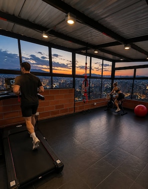 Ein Fitnessstudio mit Laufband, Ergometer und Trainingsball, mit Blick auf die Stadt durch große Fenster.