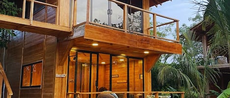A two-story wooden house with a balcony, large windows, and a porch area.