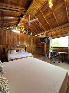 A wooden cabin room with a bed, a desk, a chair, a ceiling fan, and a television.