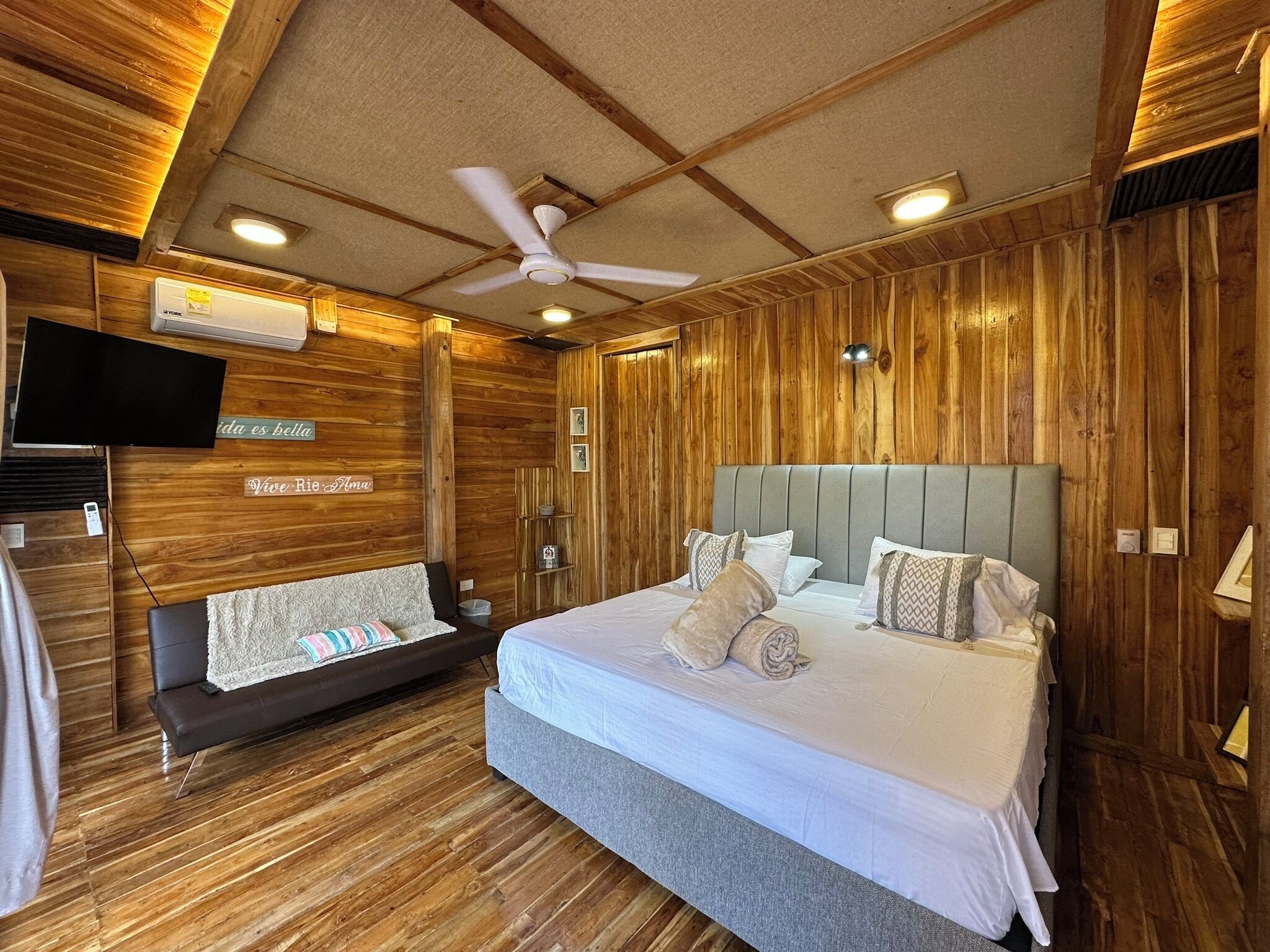A hotel room with a wooden interior, a bed with white bedding, a grey headboard, and a ceiling fan.