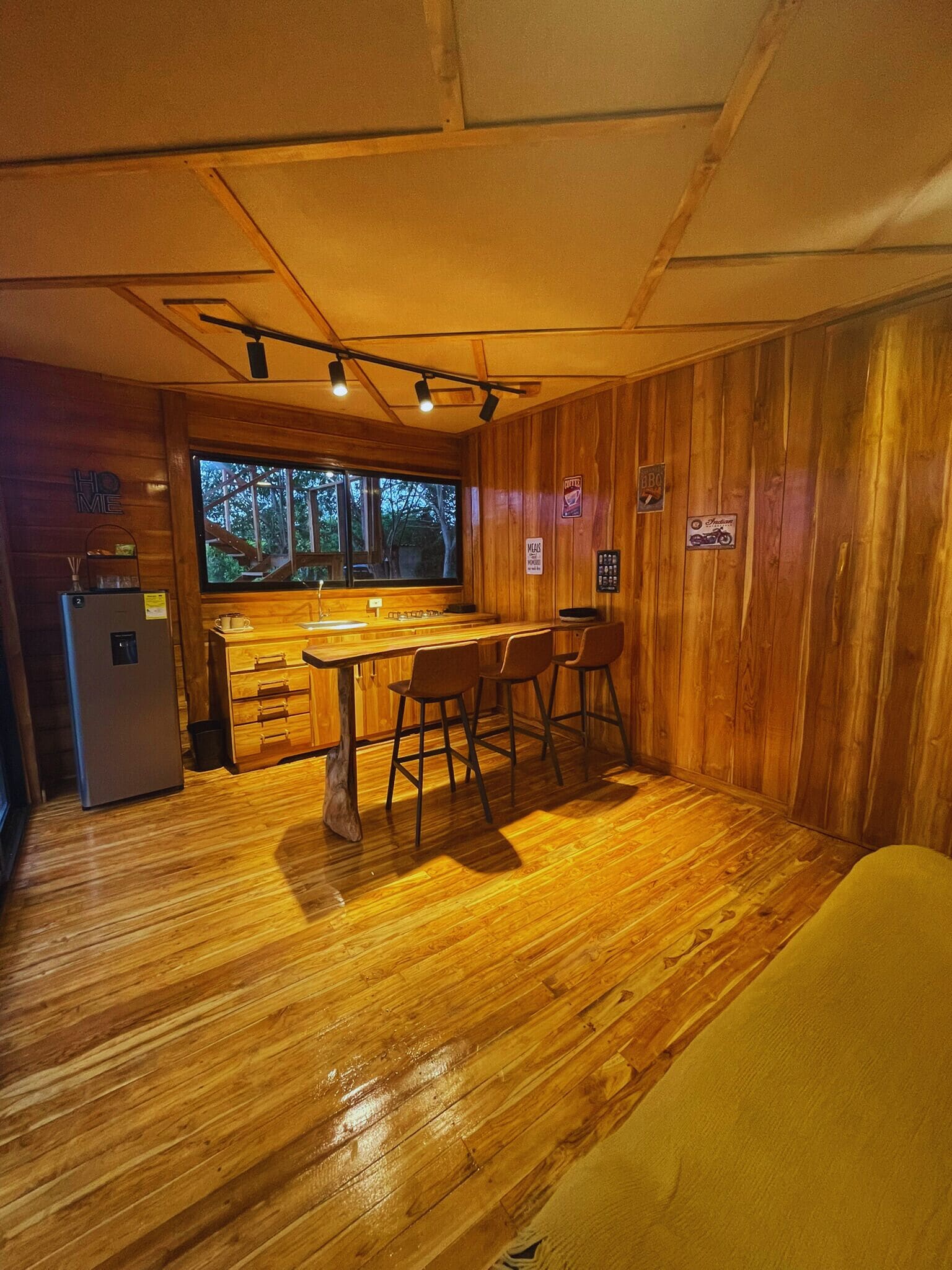 A compact kitchen with a wooden interior, a refrigerator, a sink, and a window overlooking a forest.