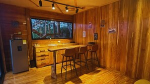 A compact kitchen with a wooden interior, a refrigerator, a sink, and a window overlooking a forest.