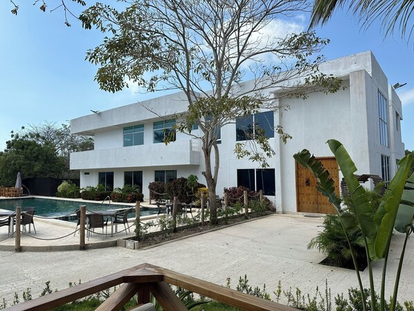 A modern two-story building with a swimming pool, outdoor seating, and tropical plants.