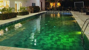 A swimming pool with a tiled deck, surrounded by a building with large windows and a tree.